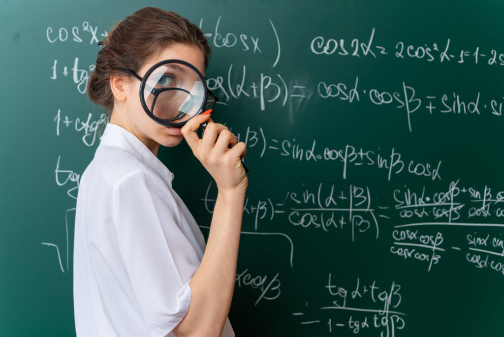 jovem-loira-confiante-professora-de-matematica-usando-oculos-de-pe-em-vista-de-perfil-na-frente-do-quadro-negro-olhando-para-a-camera-atraves-de-uma-lupa-na-sala-de-